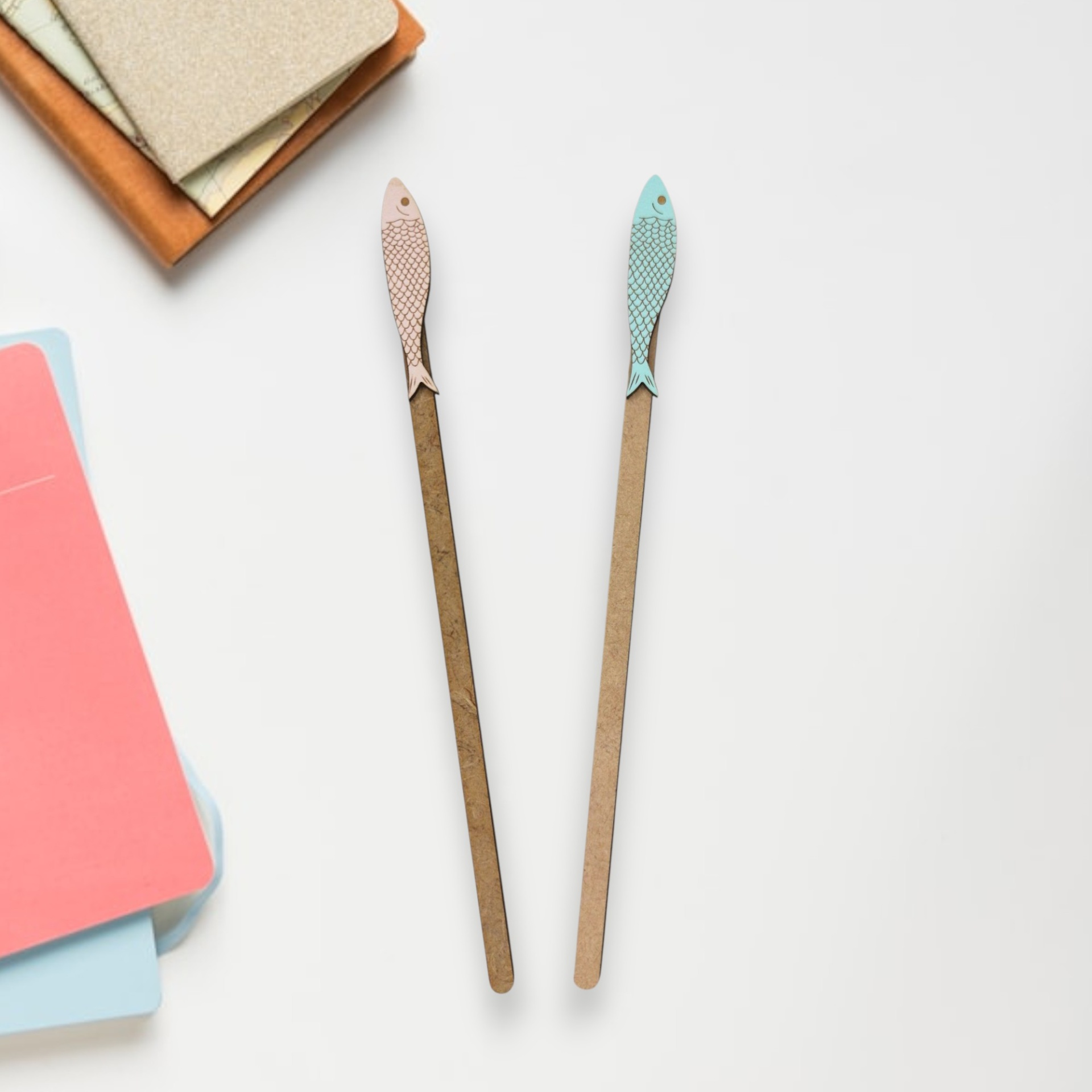 Marcador Sardinha em lata Dois lápis de madeira com pontas em forma de peixe rosa e azul, com padrão de escamas, e cadernos coloridos ao lado.