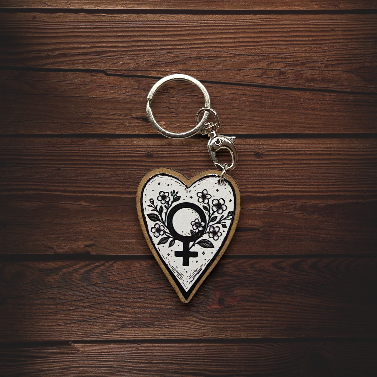 Porta-chaves Coração Mulher "Strong" Porta-chaves em forma de coração com símbolo feminino e flores em fundo branco sobre madeira castanha