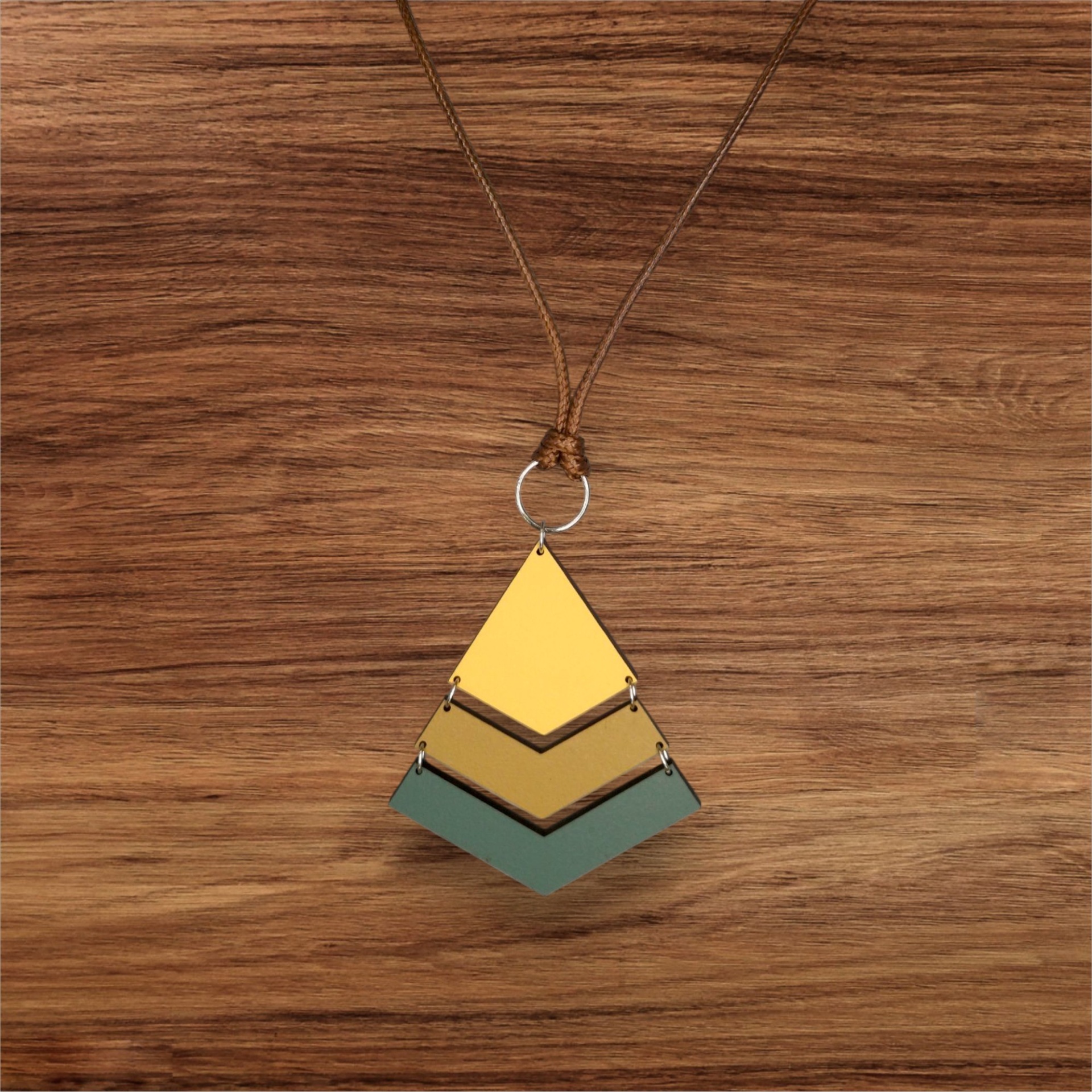 Colar Rute Colar com pendente triangular em amarelo, dourado e verde com cordão castanho sobre fundo de madeira.