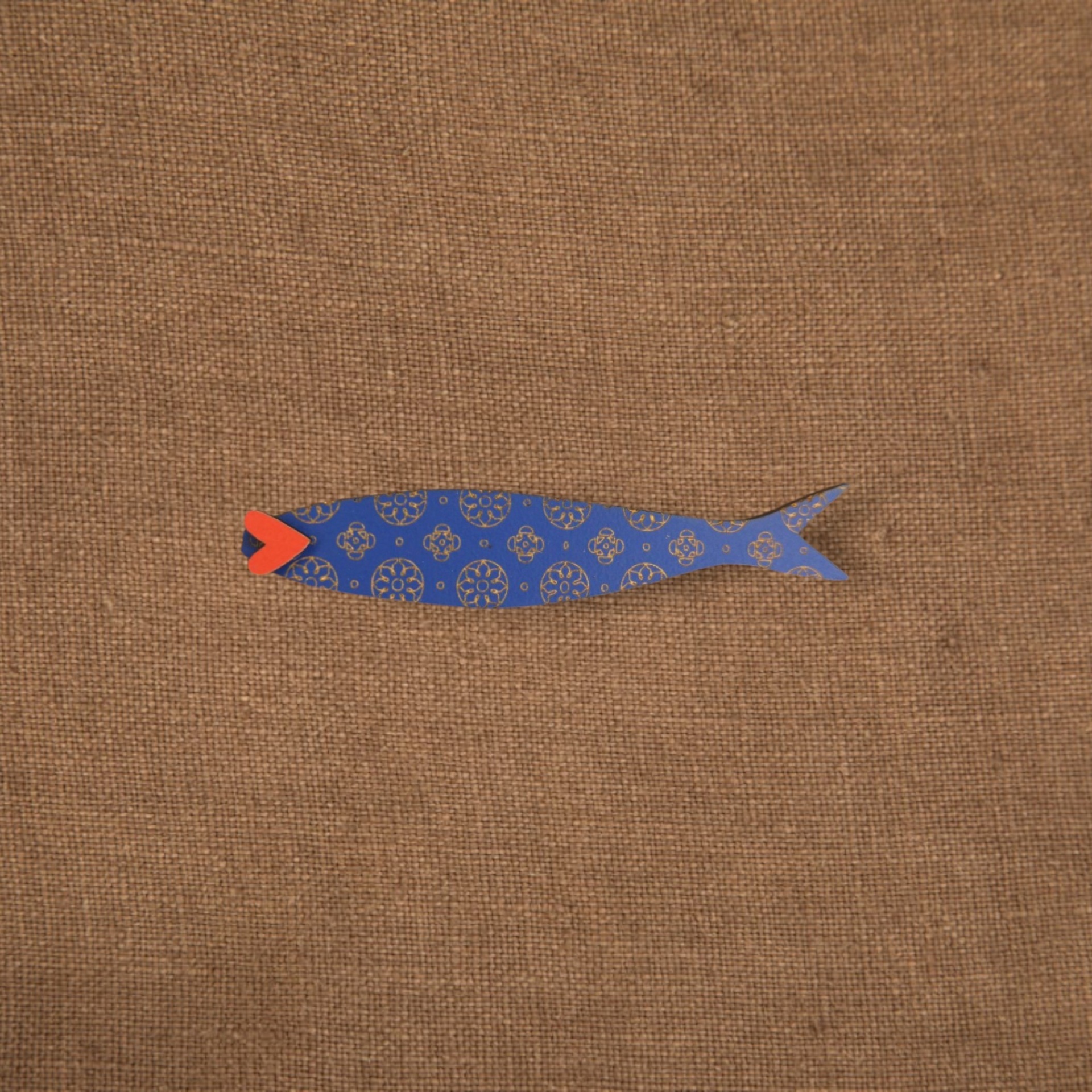 Pregadeira Sardinha Peixe decorativo azul com padrão dourado e boca vermelha sobre tecido castanho.