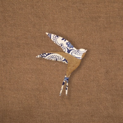 Recorte em forma de pássaro azul, branco e castanho sobre fundo de tecido bege.