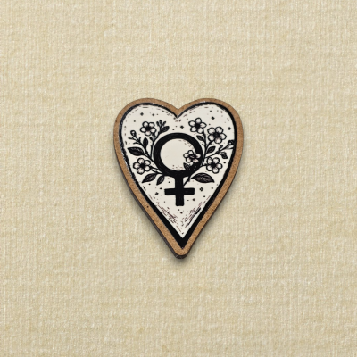 Broche coração com símbolo feminino e flores em madeira sobre fundo bege.