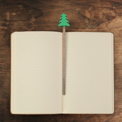 Caderno aberto com marcador de livro em forma de árvore verde sobre mesa de madeira.