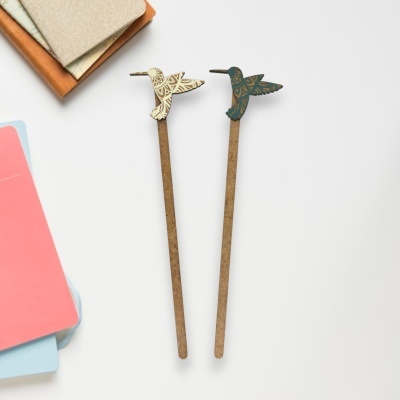 Dois lápis com decoração de beija-flor em bege e verde sobre uma mesa branca com cadernos coloridos ao lado