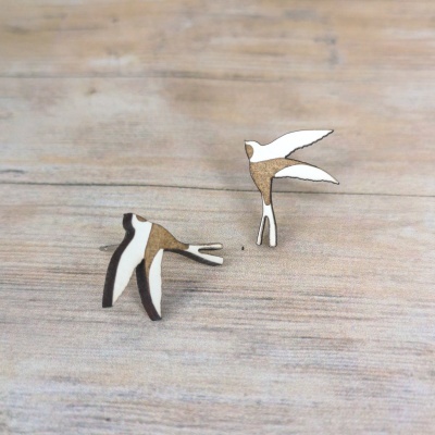 Brincos em forma de andorinha de madeira branca e castanha sobre superfície de madeira clara