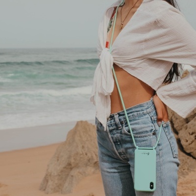 Telemóvel com capa verde-água e cordão CASetiFY, mulher com blusa branca e jeans na praia