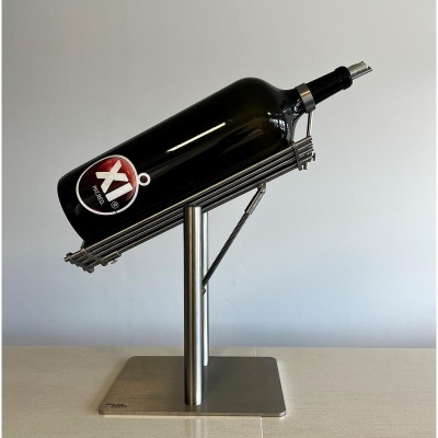 Garrafa de vidro escuro com rótulo XI ÁRIDO num suporte metálico prateado.