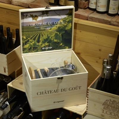 Caixa de madeira aberta com utensílios para vinho, rodeada por garrafas e caixas em adega