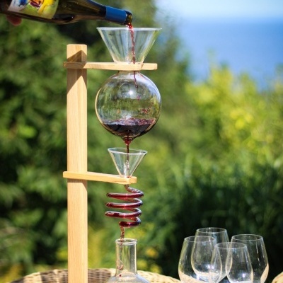 Dispositivo de vidro para aerar vinho com suporte de madeira e quatro copos de vinho vazios ao lado numa mesa ao ar livre