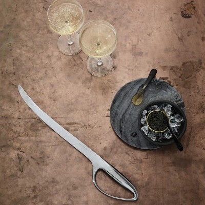 Dois copos de champanhe, faca metálica longa, recipiente preto com conteúdo em base cinza escura numa mesa de madeira.