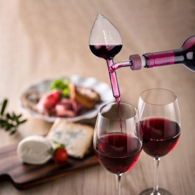 Decantador de vinho transparente a servir vinho tinto em taças com queijos e frios ao fundo
