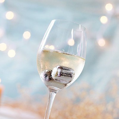 Copo de vinho com líquido branco e cubos de gelo metálicos sobre fundo azul claro com luzes desfocadas