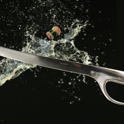 Faca curva em aço inoxidável com cabo aberto em fundo preto, garrafa de champanhe e líquido salpicando