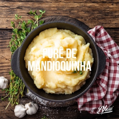 Purê de mandioquinha numa panela preta com alho, ervas frescas e pano às riscas numa tábua de madeira