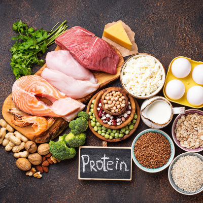 Alimentos ricos em proteína como carne, peixe, ovos, queijo, legumes, nozes e uma tábua com a palavra 'Protein'