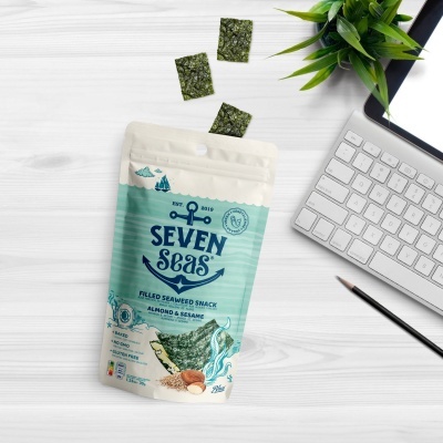 Pacote de petisco de algas Seven Seas e três pedaços de alga numa mesa branca com teclado, caderno e planta