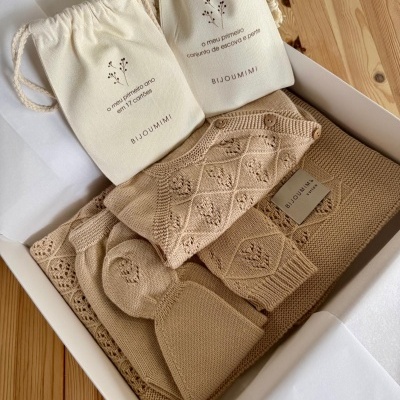 Conjunto de roupa de bebé bege em malha com padrão e botões e dois sacos de tecido branco com texto, dentro de caixa branca