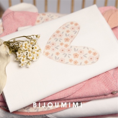 Toalha branca com coração floral sobre tecidos rosa e ramo de flores brancas.