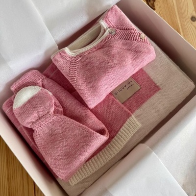Conjunto de roupa de bebé rosa e branca em caixa branca sobre superfície de madeira.