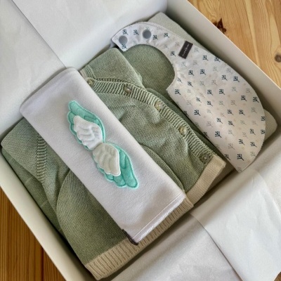 Caixa aberta com roupa de bebé verde, bandana branca estampada e pano branco com asas azuis e brancas