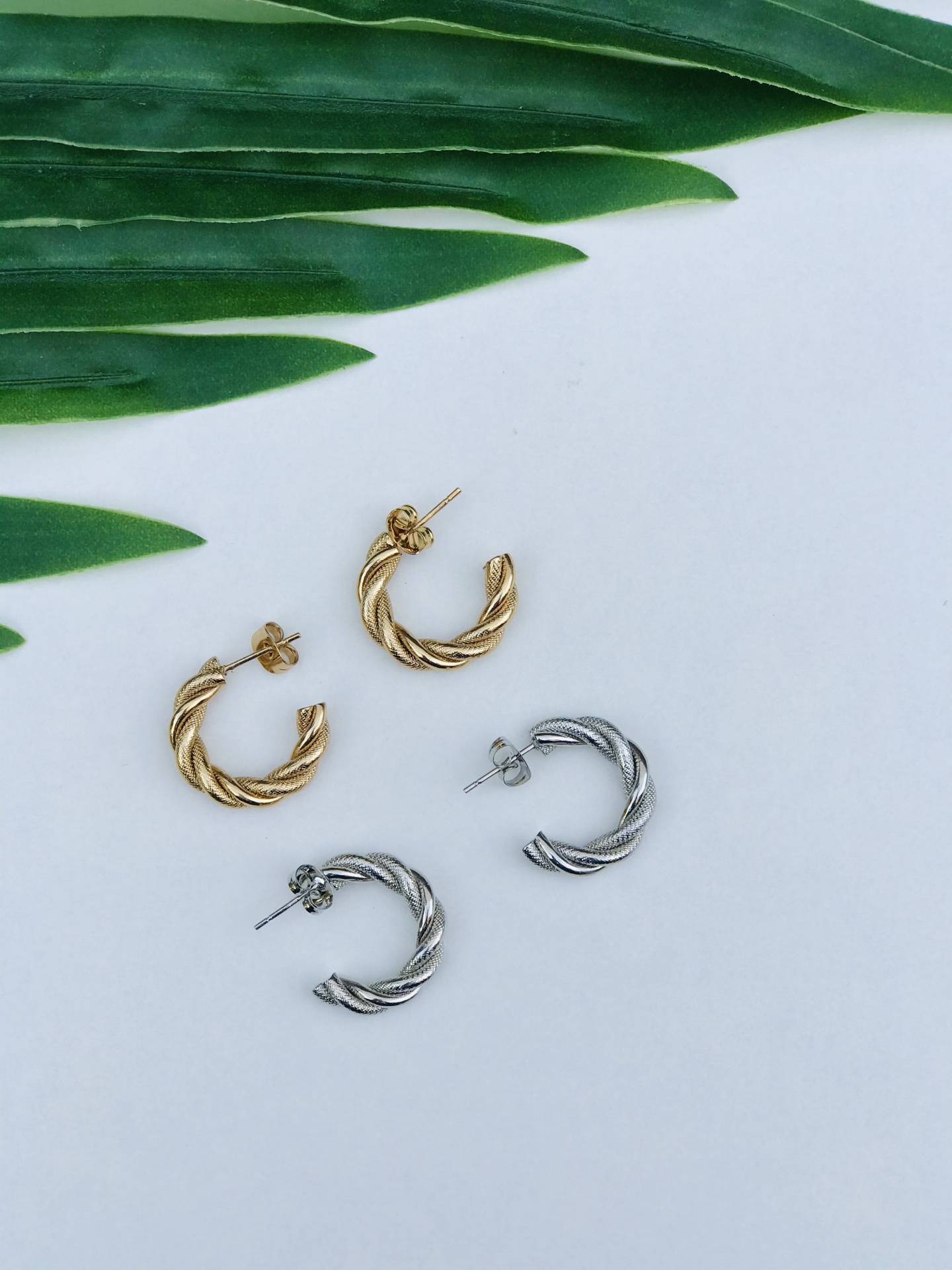 Dois pares de brincos argola torcida dourado e prateado com folhas verdes ao fundo
