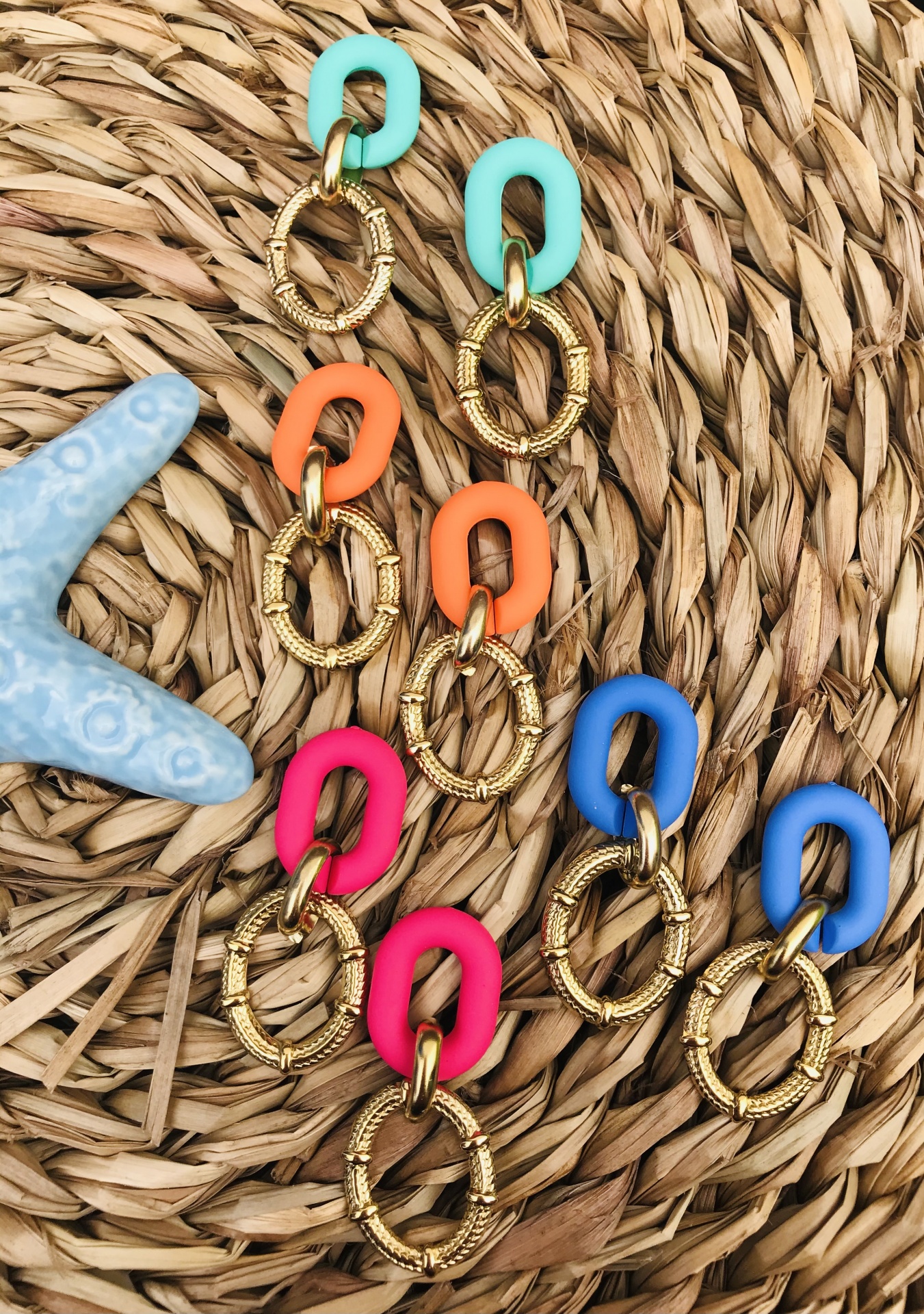 Brincos coloridos com argolas de resina e douradas sobre tapete de palha com estrela do mar azul