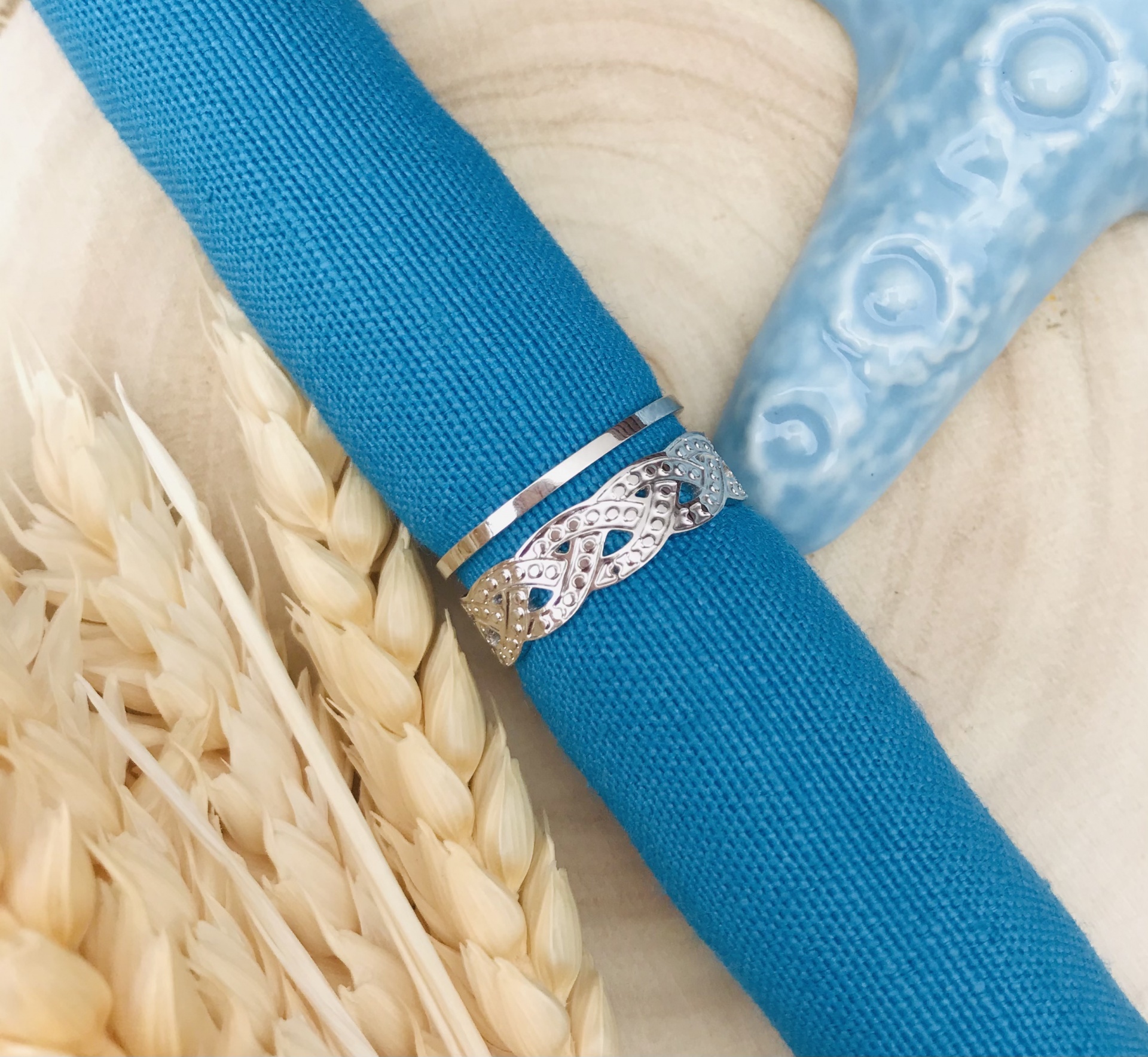 Anel prateado entrelaçado com pedras num guardanapo azul sobre madeira clara com espigas de trigo e objeto azul
