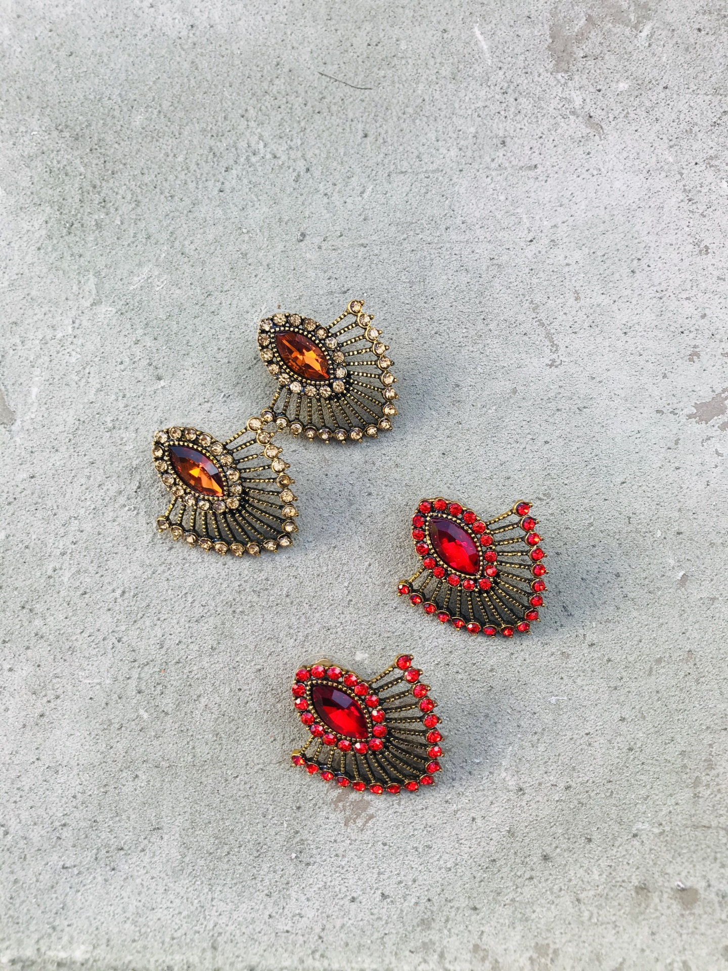 Quatro brincos em forma de folha com pedras coloridas e detalhes dourados ou vermelhos sobre fundo cinzento.