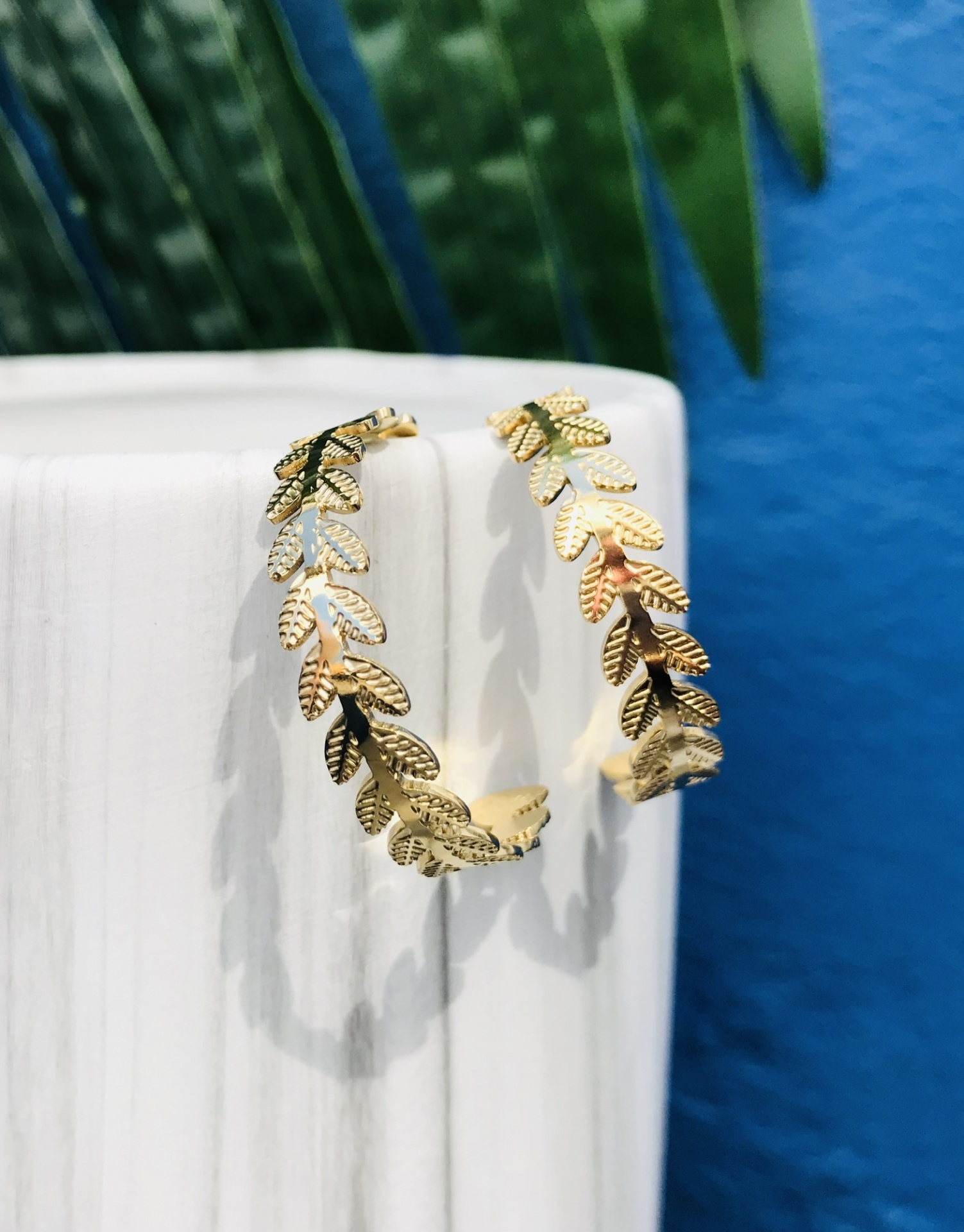 Brincos dourados em padrão de folhas numa superfície branca com fundo azul e folhas verdes.