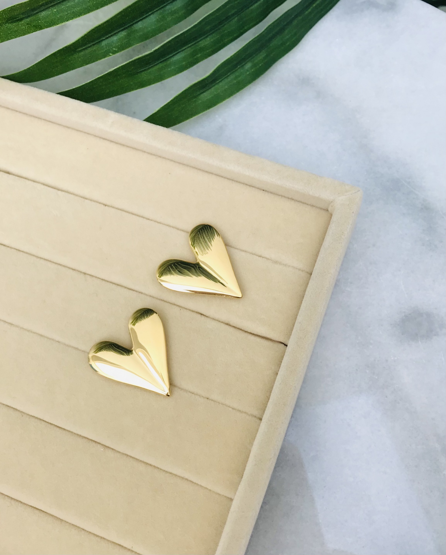 Brincos de coração dourados sobre base de veludo bege com folhas verdes ao fundo