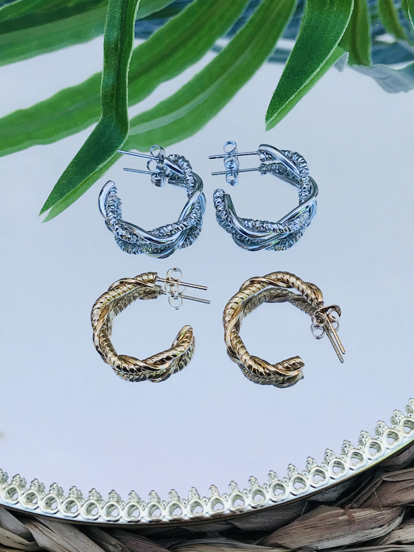 Dois pares de brincos argola trançada prateada e dourada sobre espelho com folhas verdes ao fundo