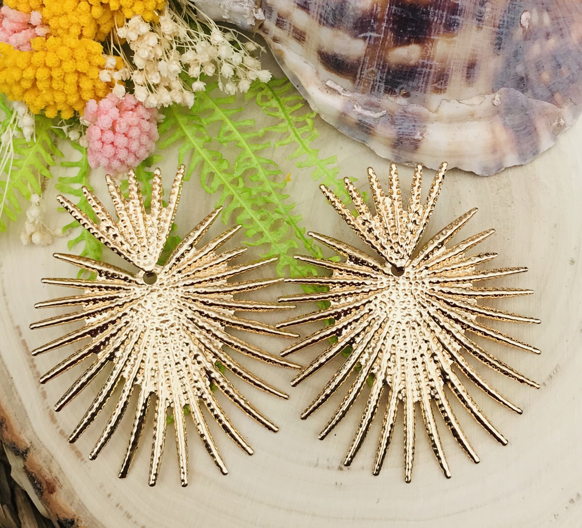 Brincos dourados com design de folhas secas, sobre madeira com conchas e flores artificiais.