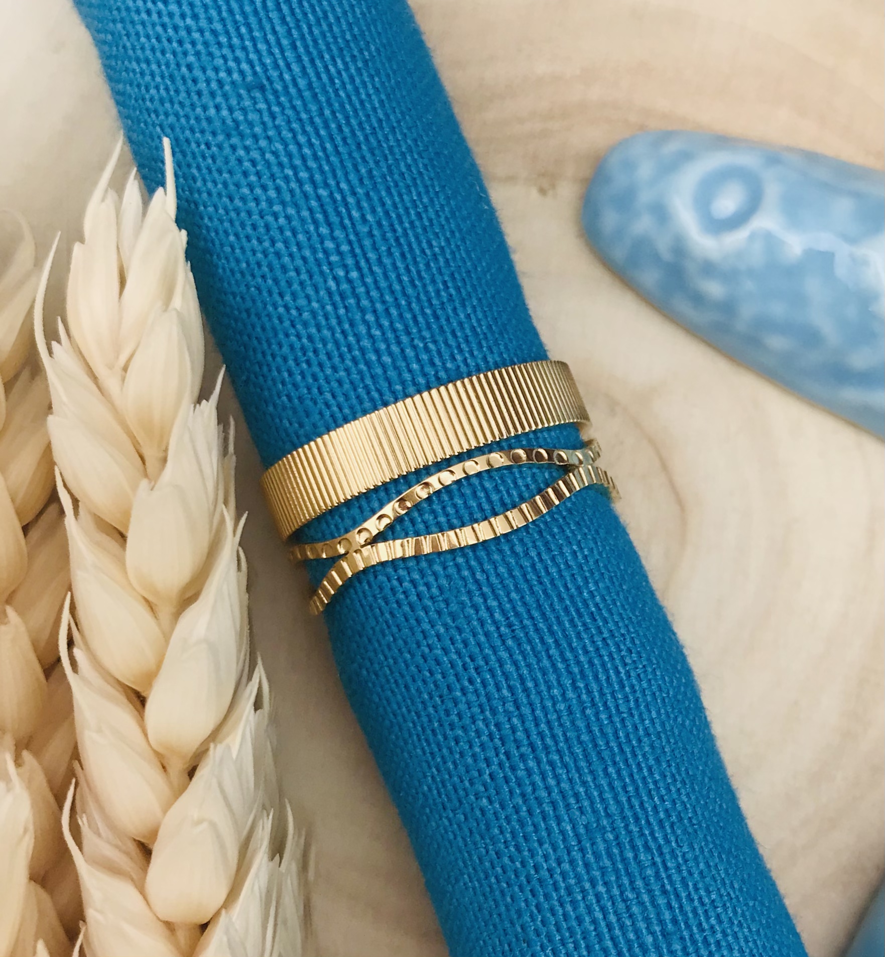Anel dourado de três bandas sobre tecido azul com espigas secas ao lado