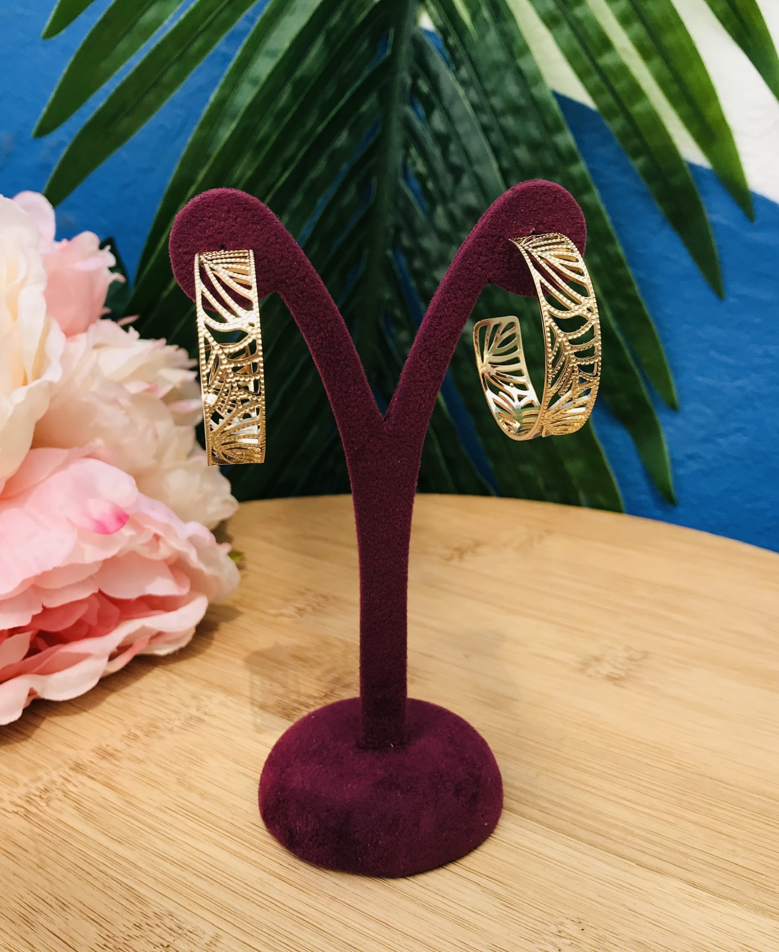 Brincos dourados em suporte de veludo roxo com flores e folhas ao fundo