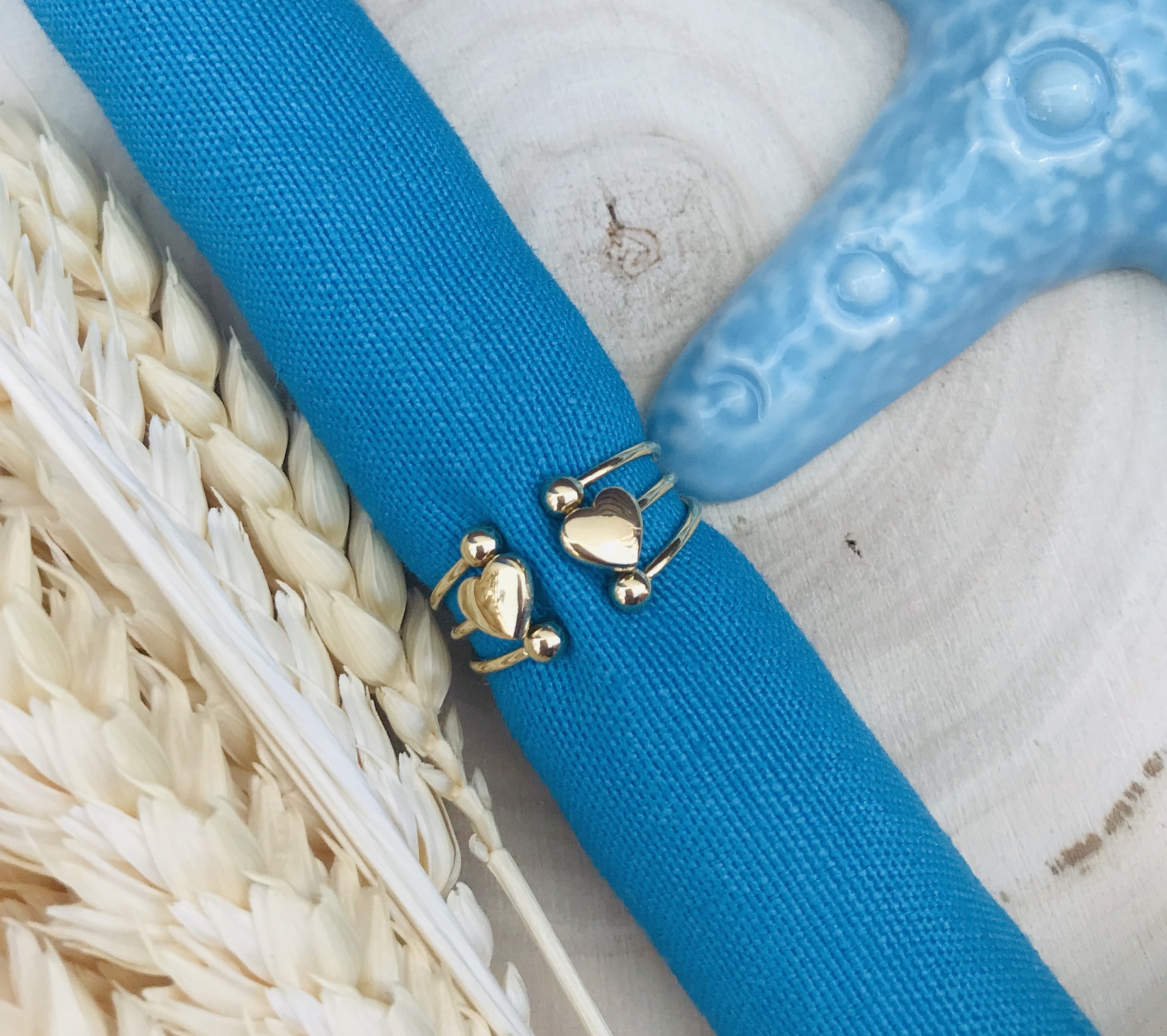 Anéis dourados com coração sobre pano azul em madeira clara com decoração de estrela do mar e trigo.