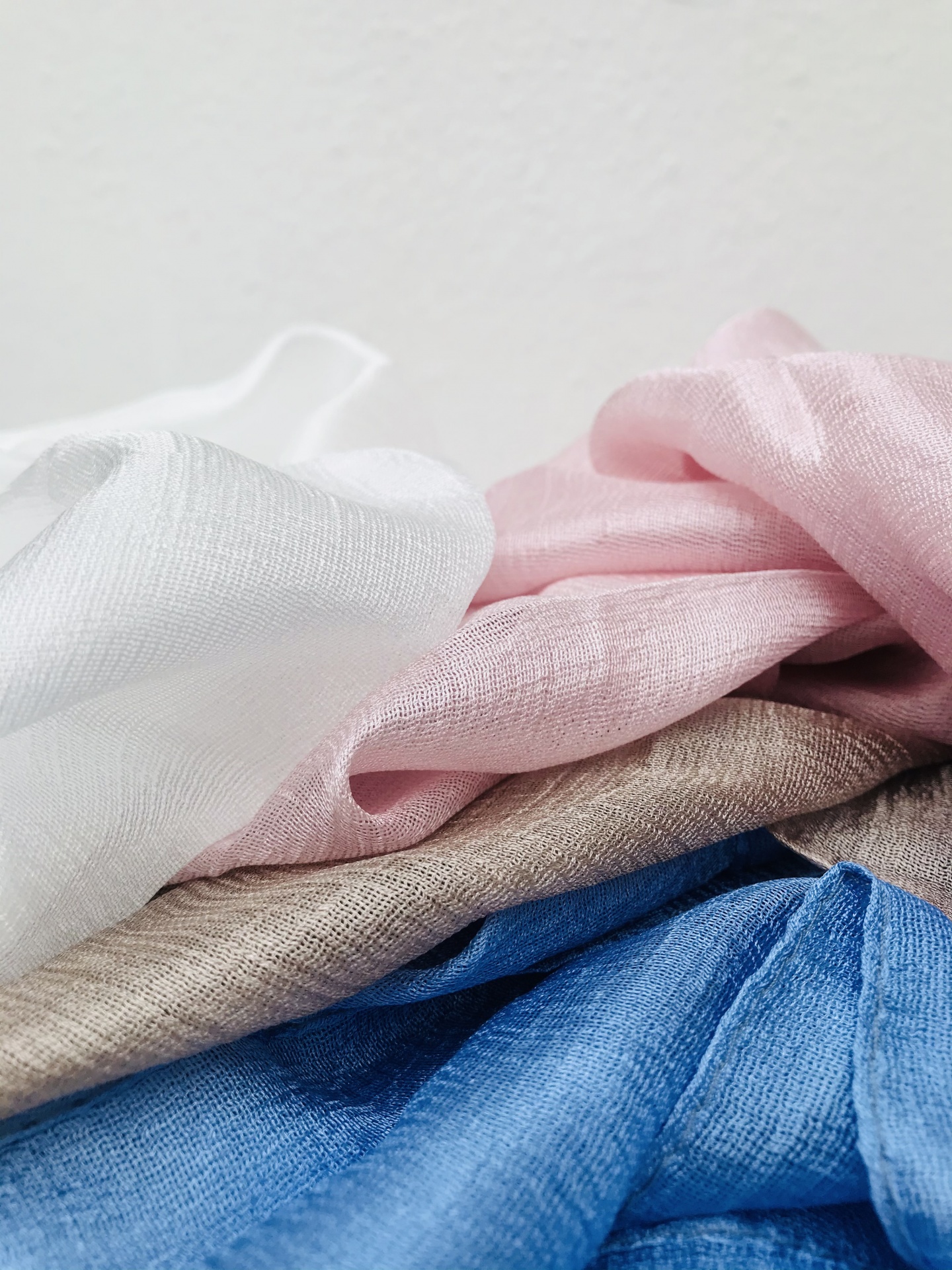 Tecidos finos em branco, rosa, bege e azul dispostos em camadas onduladas com fundo claro.