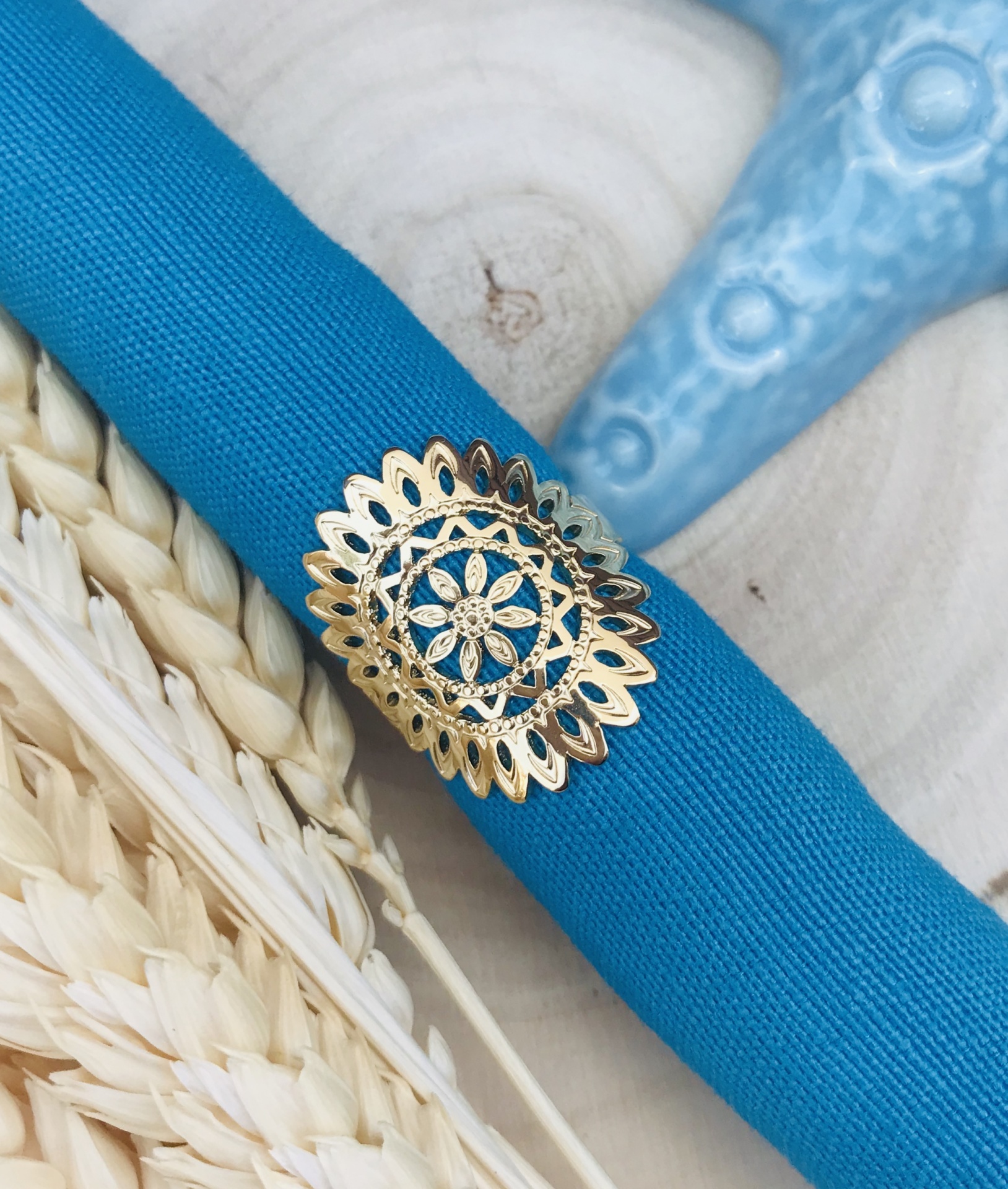 Guardanapo azul com anel dourado com padrão floral sobre mesa de madeira clara com decoração azul e palha