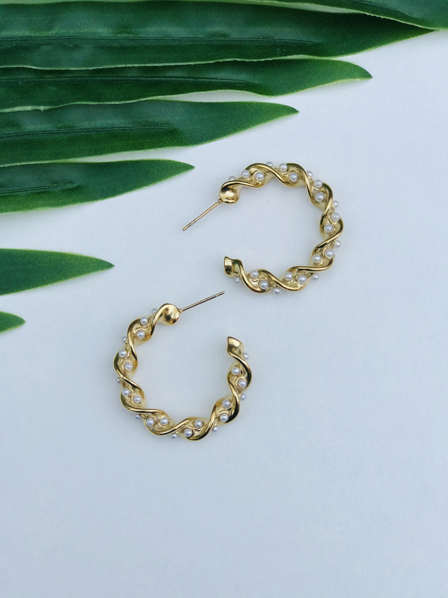 Brincos de argola dourados entrelaçados com pérolas sobre fundo branco e folhas verdes