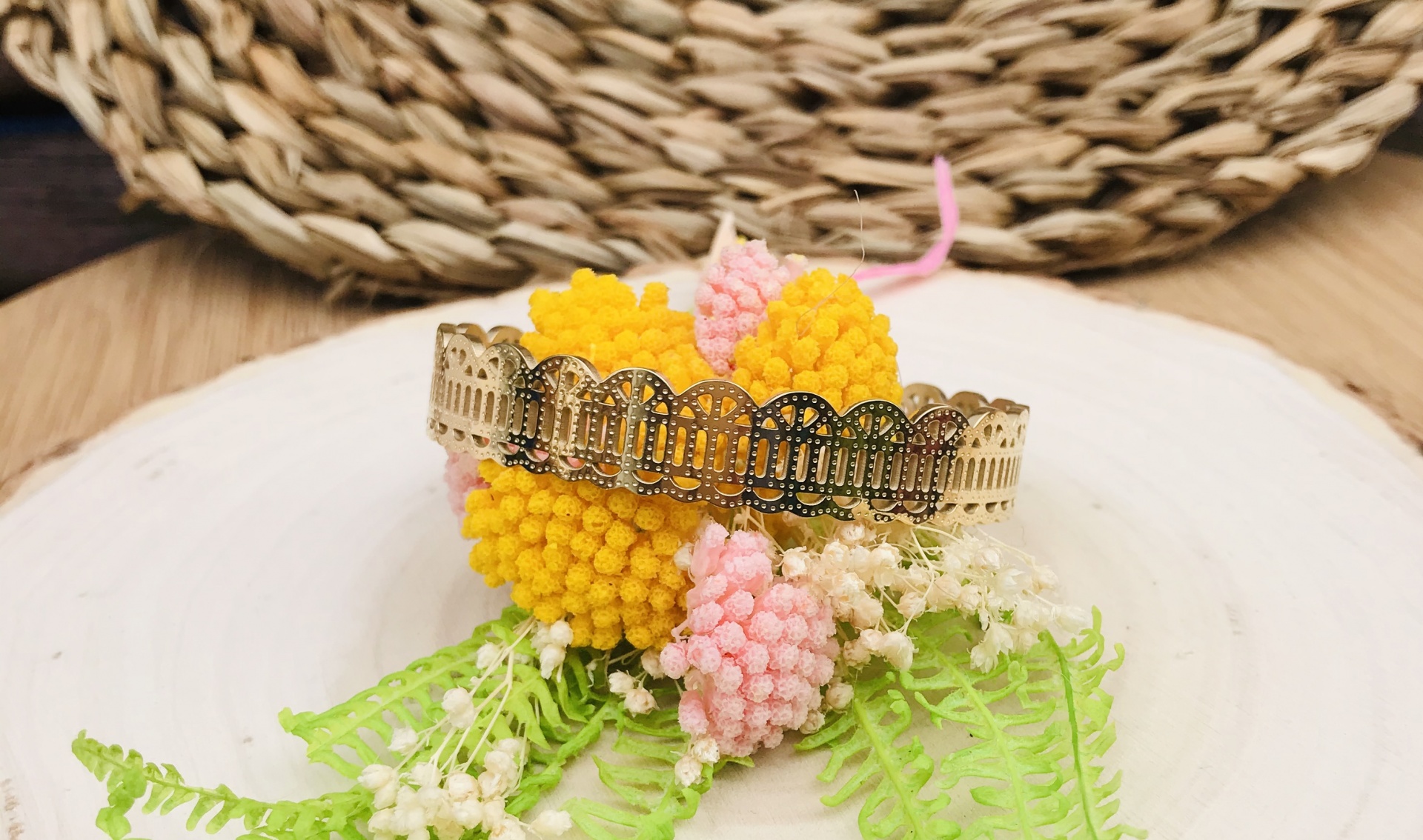 Pulseira dourada com padrão recortado sobre flores decorativas amarelas e cor-de-rosa com folhas, em fundo de madeira e tapete de palha.