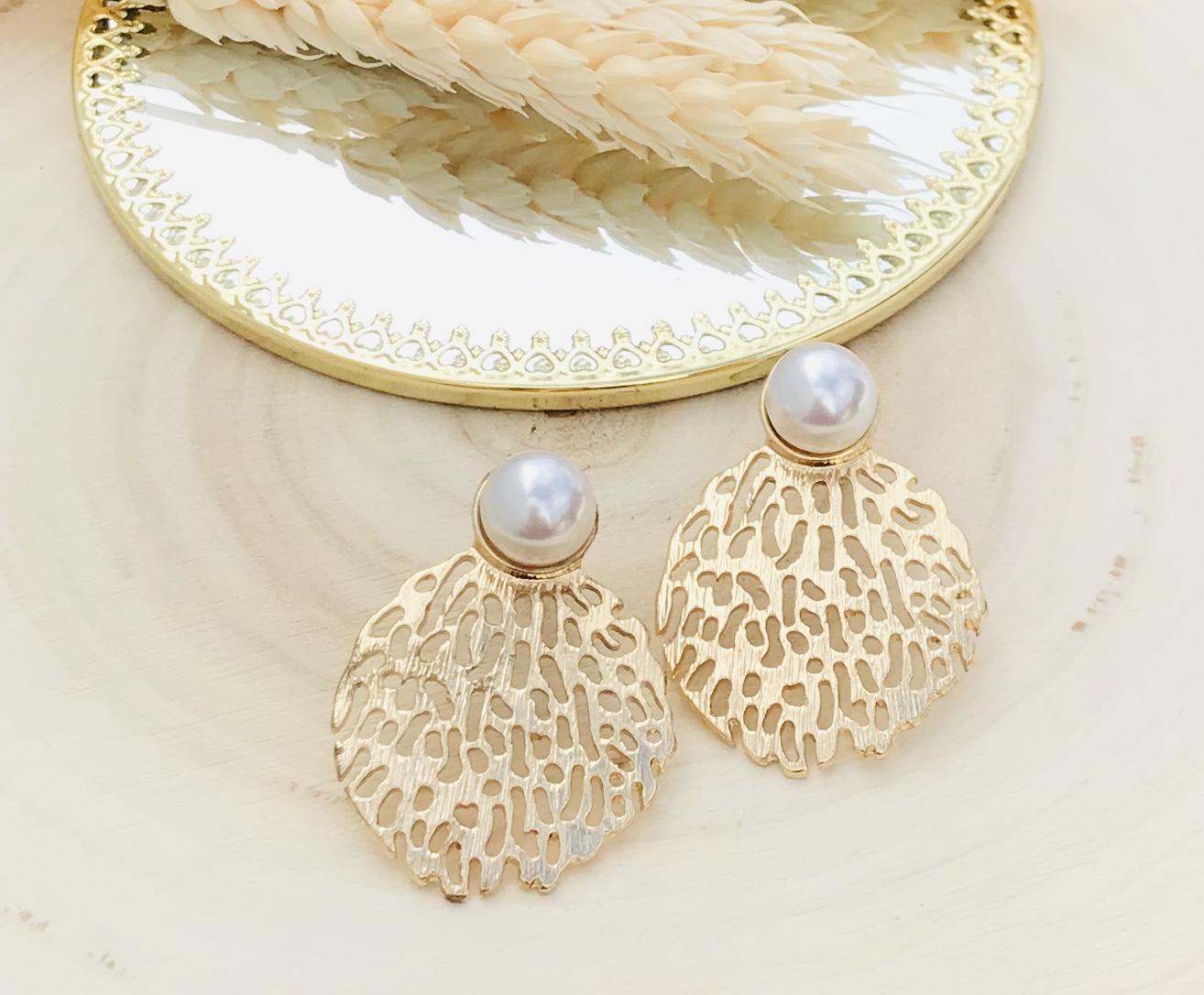 Brincos dourados com pérola branca sobre prato espelhado e superfície clara