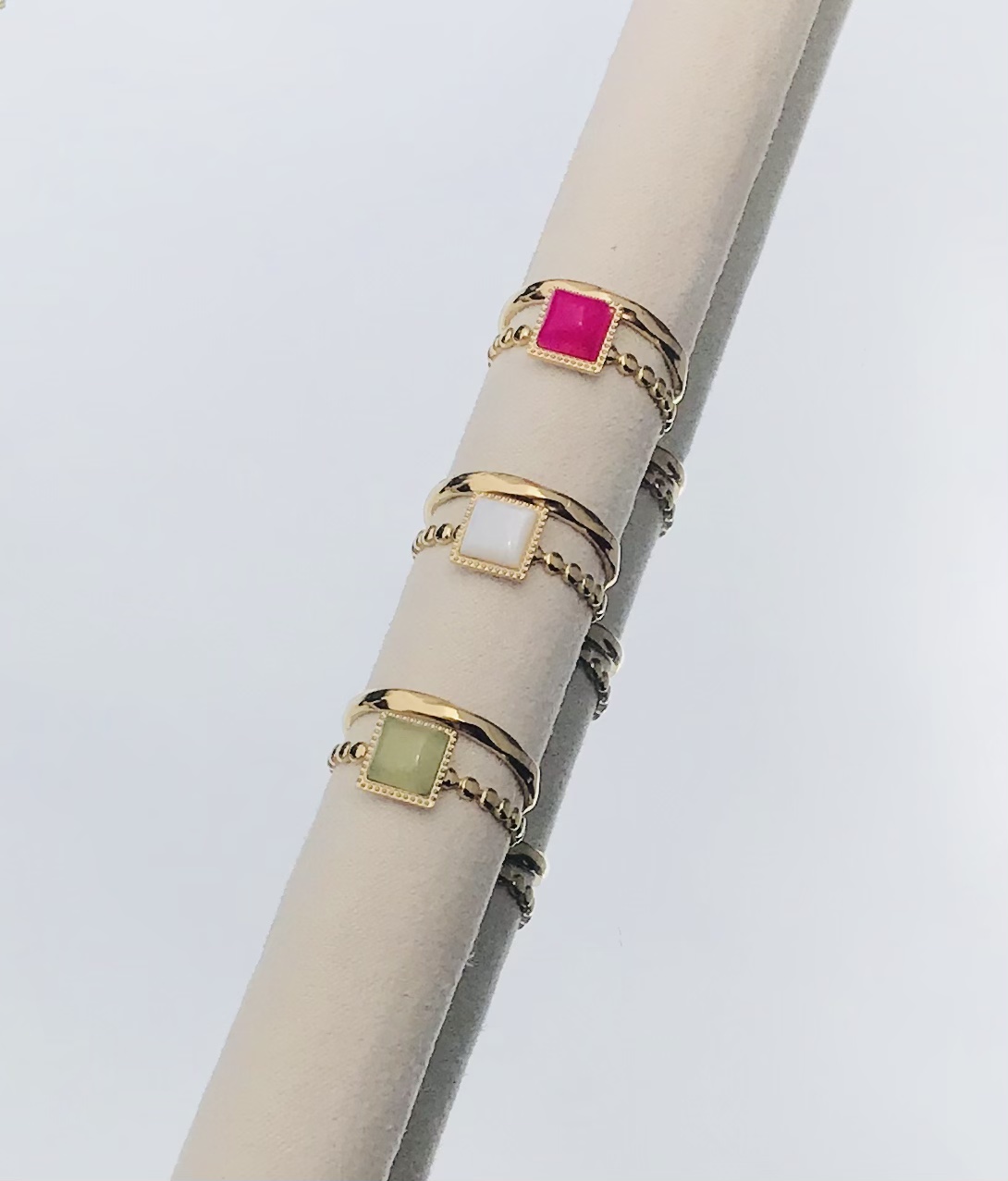 Três anéis dourados com pedras quadradas rosa, branco e verde num suporte bege