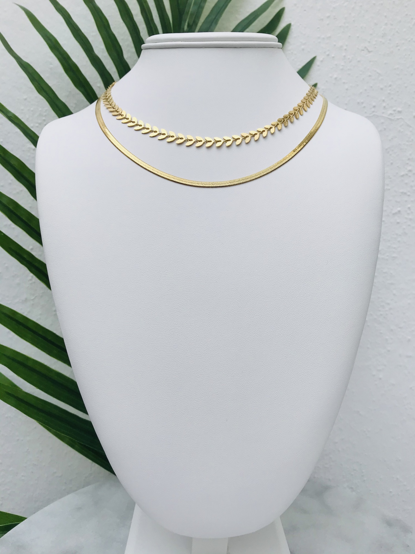 Duas gargantilhas douradas num busto branco com folhas verdes ao fundo