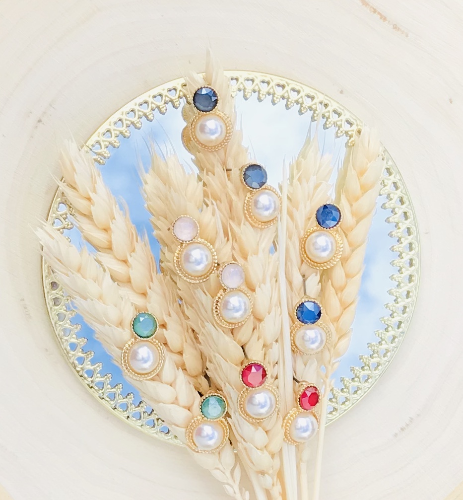 Brincos dourados com pérolas e pedras coloridas sobre espigas de trigo secas em espelho.