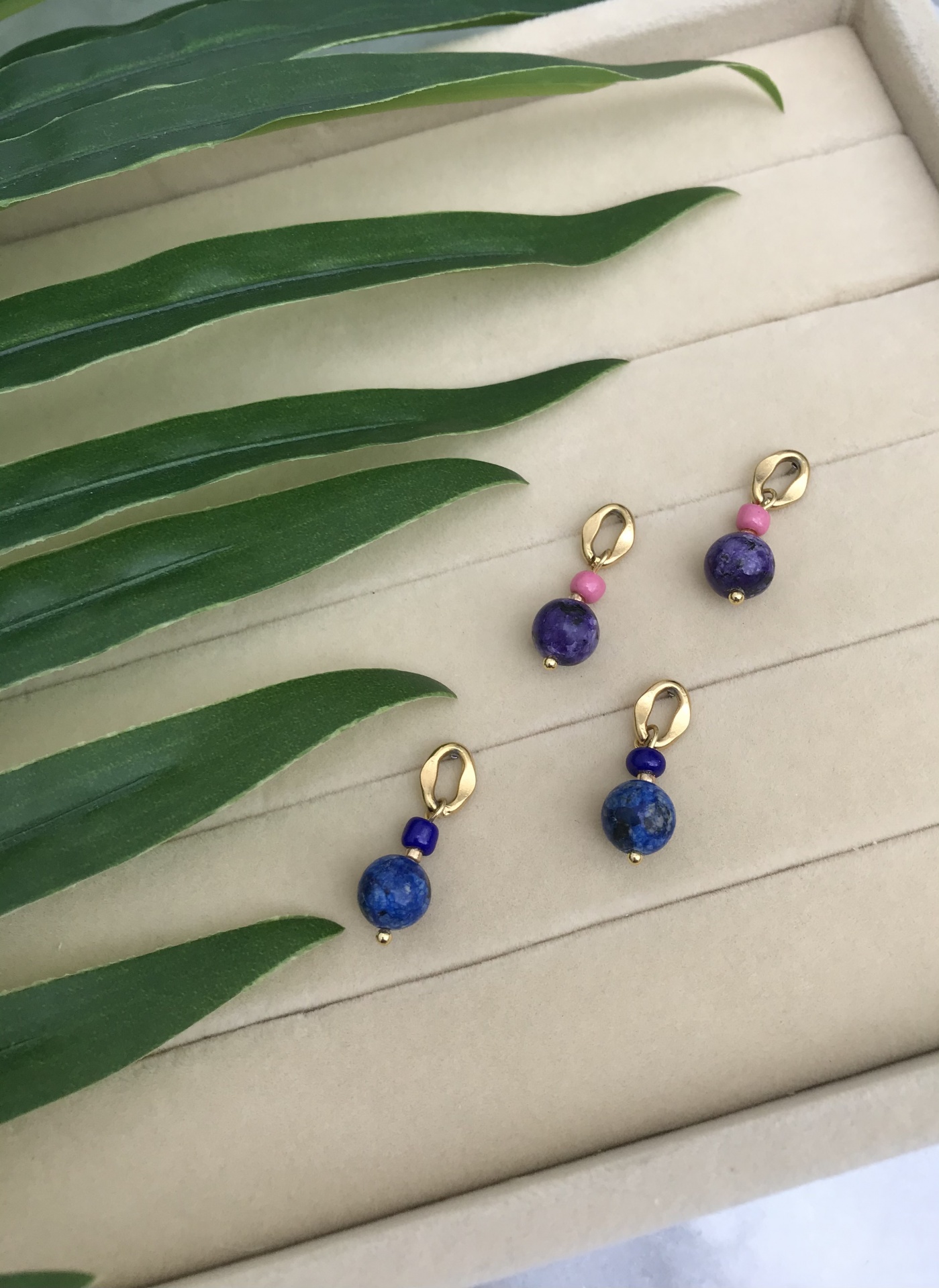 Dois pares de brincos com contas coloridas e fecho dourado sobre fundo de tecido bege e folhas verdes