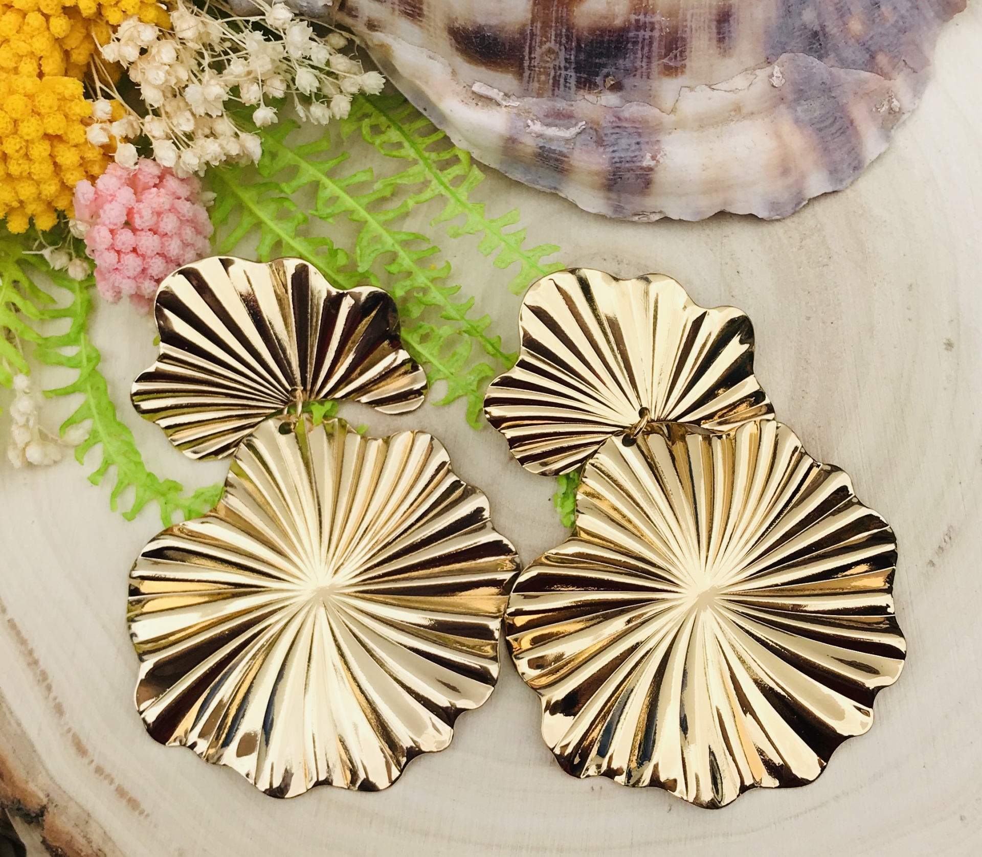 Brincos dourados em forma de folhas com relevo em fundo claro com flores e concha