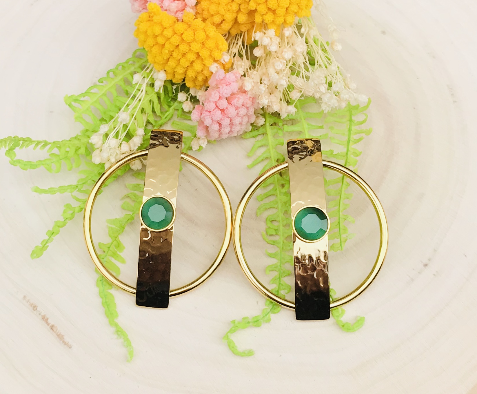 Brincos dourados com pedras verdes e aro circular, sobre base branca com flores decorativas