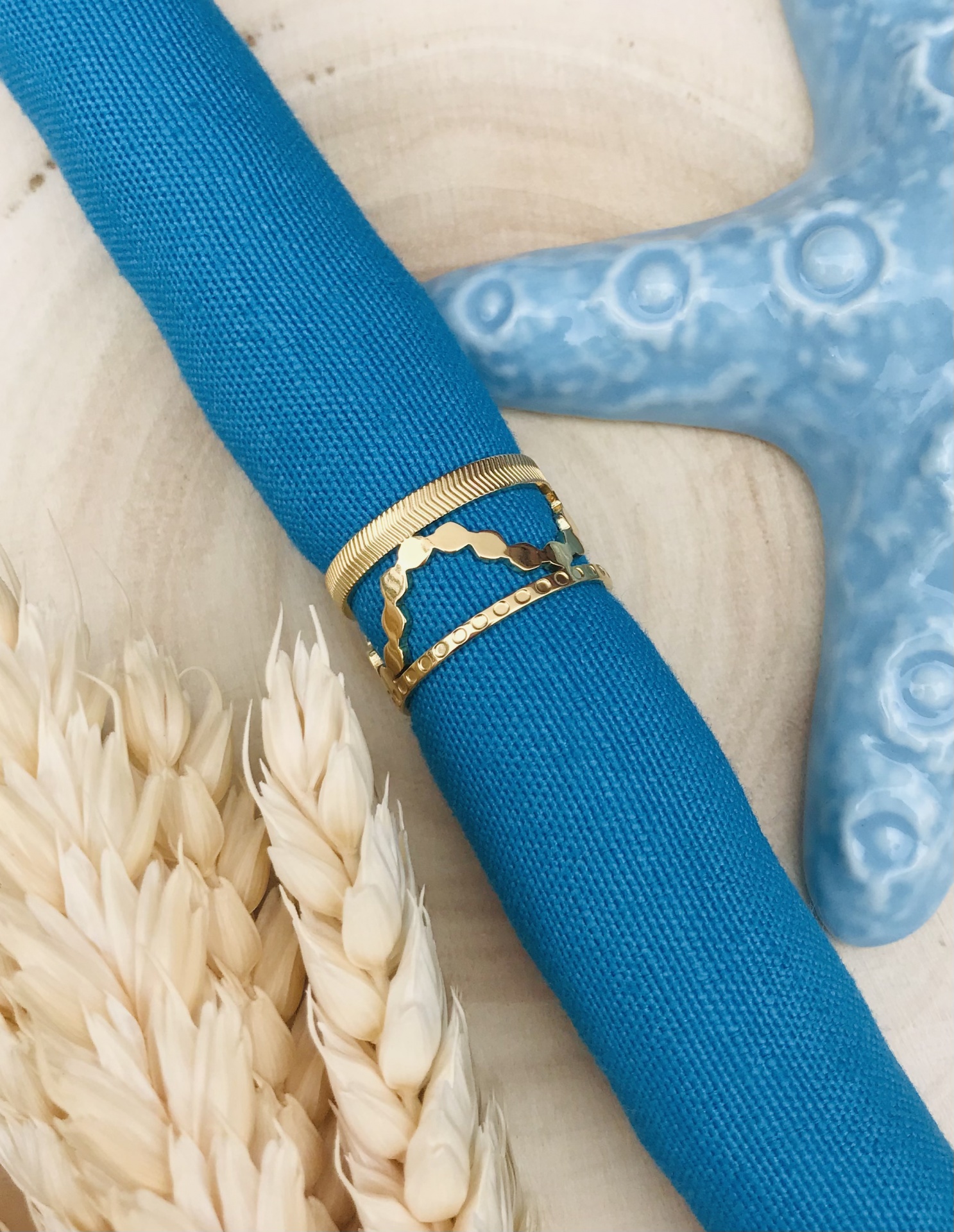 Anel dourado com três faixas sobre tecido azul com trigo seco e estrela-do-mar azul
