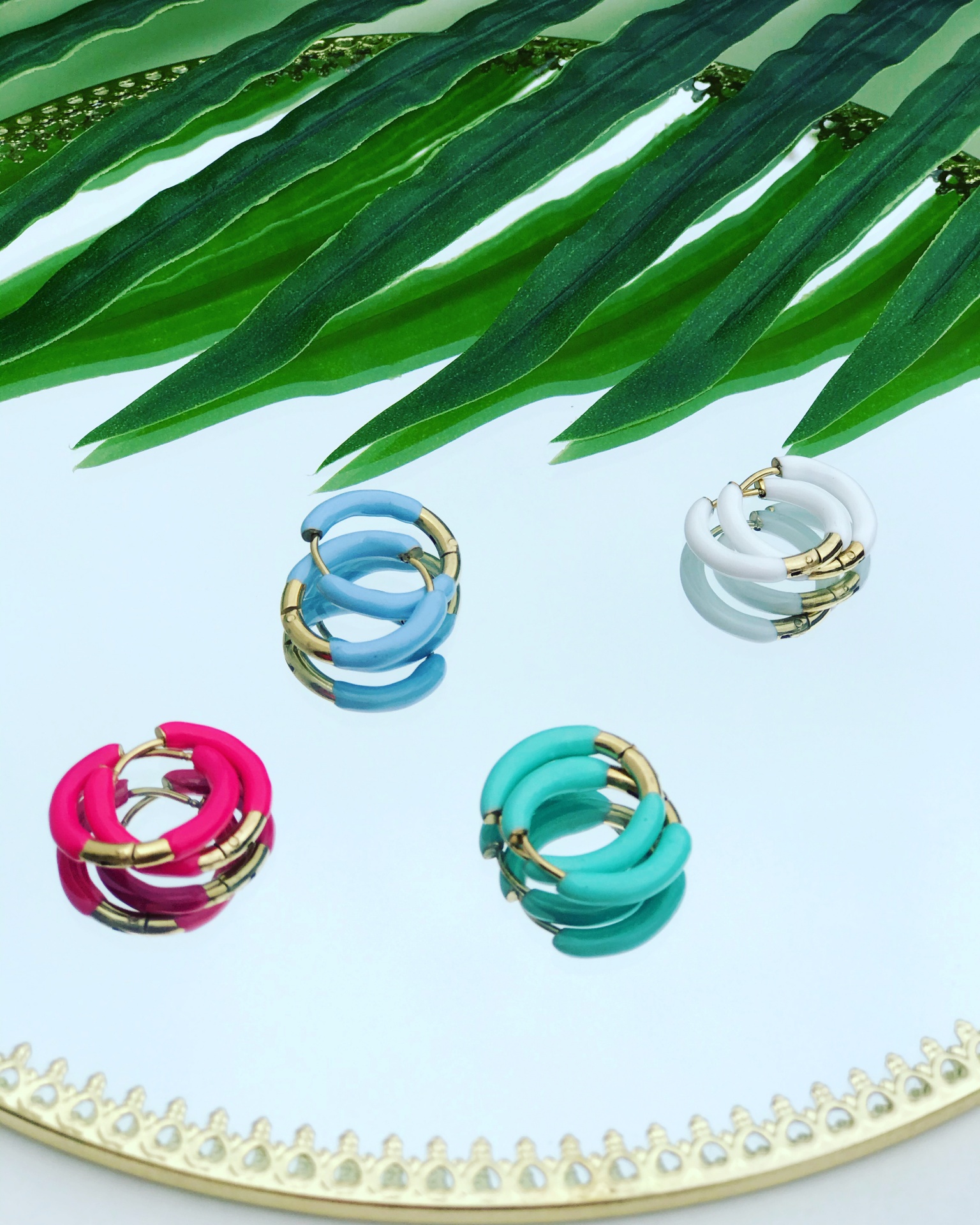 Quatro pares de brincos coloridos em argola com detalhes dourados sobre um espelho redondo com borda dourada, com folhas verdes ao fundo.