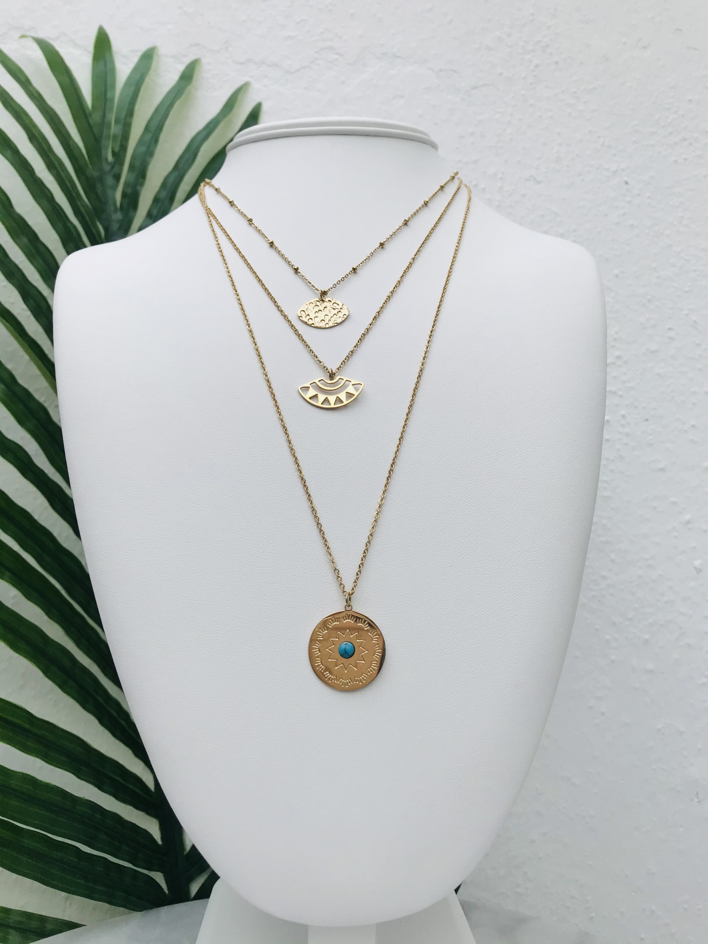 Colar triplo dourado com três pendentes em busto branco e folhas verdes ao fundo