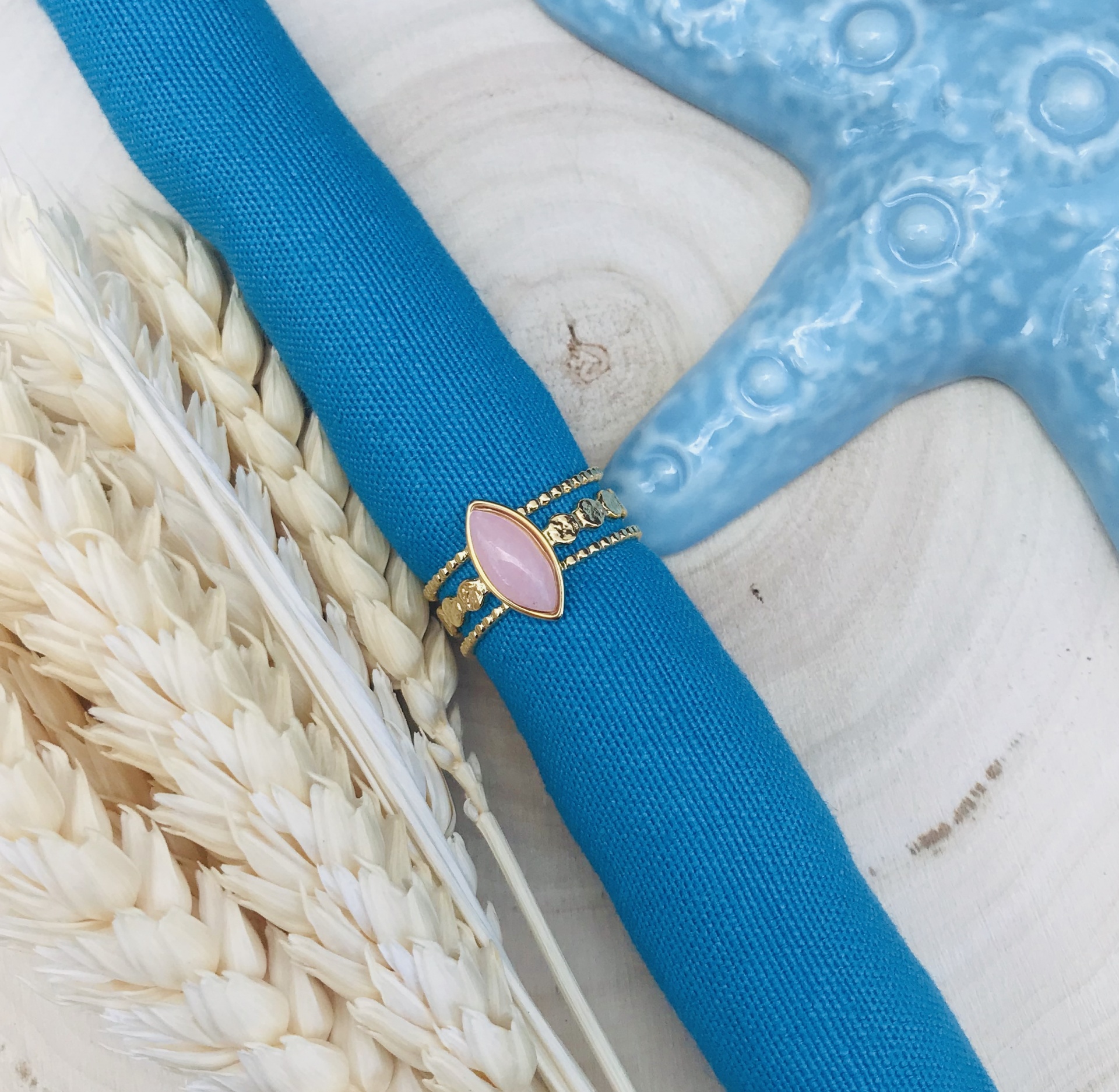 Anel dourado com pedra rosa sobre tecido azul, espigas de trigo secas e estrela do mar azul sobre fundo de madeira.